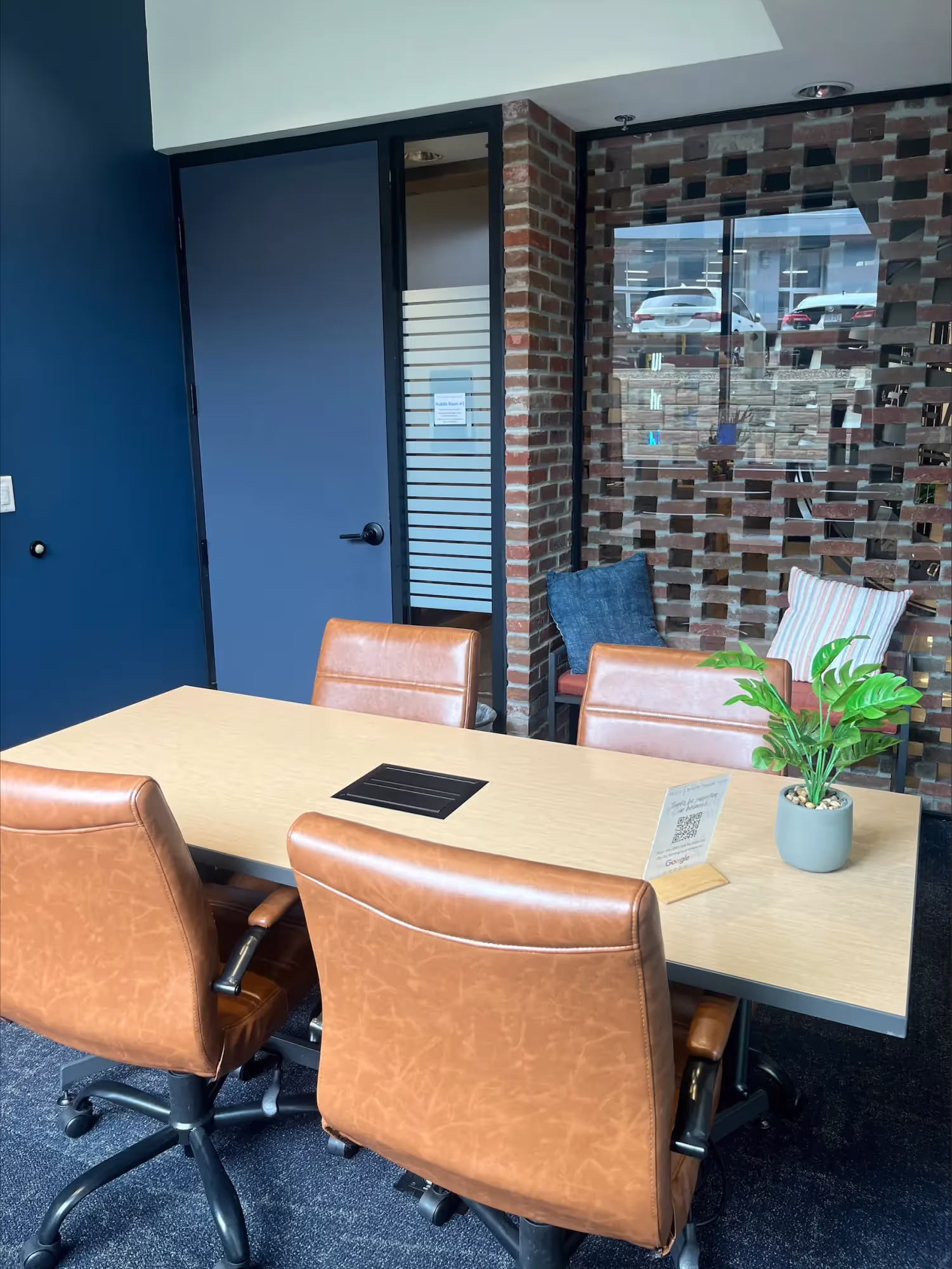 A modern coworking space meeting room with a wooden table, three brown leather chairs, a potted plant, and a blue wall. A glass wall with a brick pattern and a window showing parked cars are in the background.