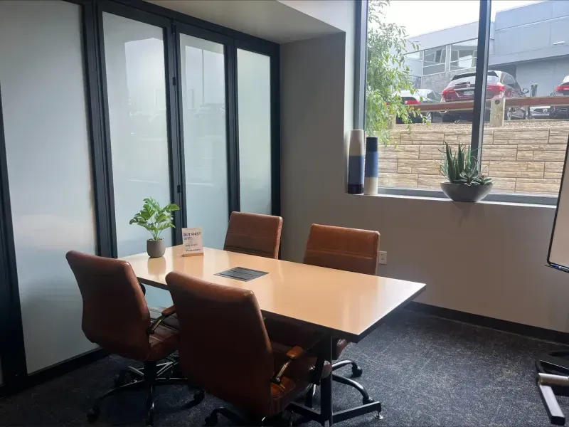 A modern meeting room with four brown chairs around a rectangular white table, a small plant centerpiece, large windows letting in natural light, and outdoor cars visible—perfect for productive meetings or as part of event spaces Columbus Ohio offers.