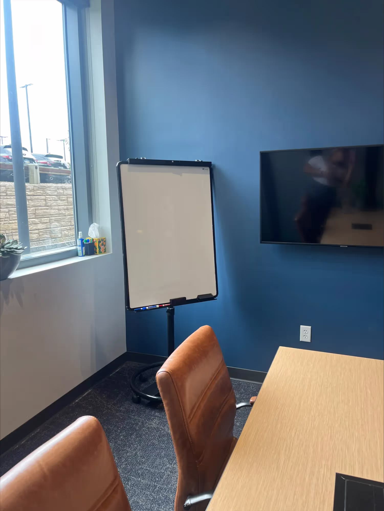 A modern conference room in an office space for rent, featuring brown chairs, a wooden table, a blank whiteboard, a flat-screen TV on a blue wall, sunlight from a window, and a small potted plant on the sill.