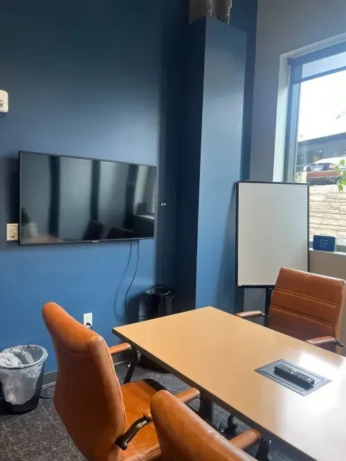 A modern coworking space meeting room with a wooden table, four brown leather chairs, a wall-mounted TV, a whiteboard, a trash can, and large windows letting in natural light. Perfect for teams needing a virtual business address.