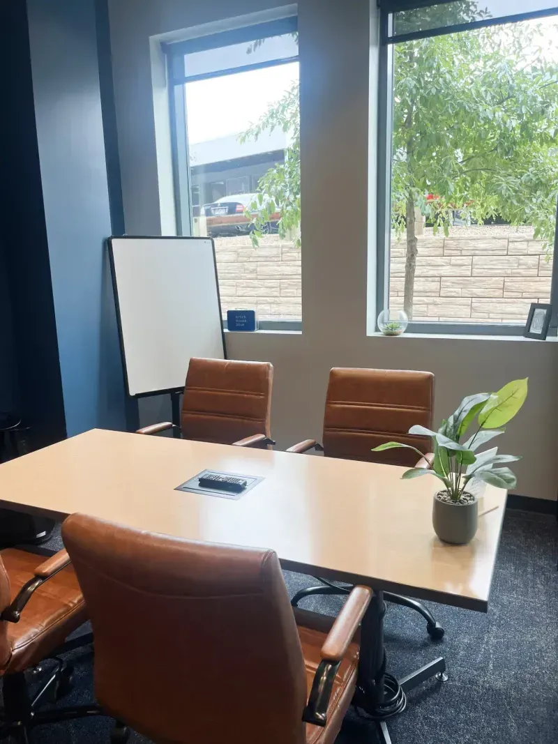 A modern conference room in a coworking space with four brown leather chairs around a rectangular table, a potted plant, a whiteboard in the corner, and two large windows filling the meeting room with natural light.