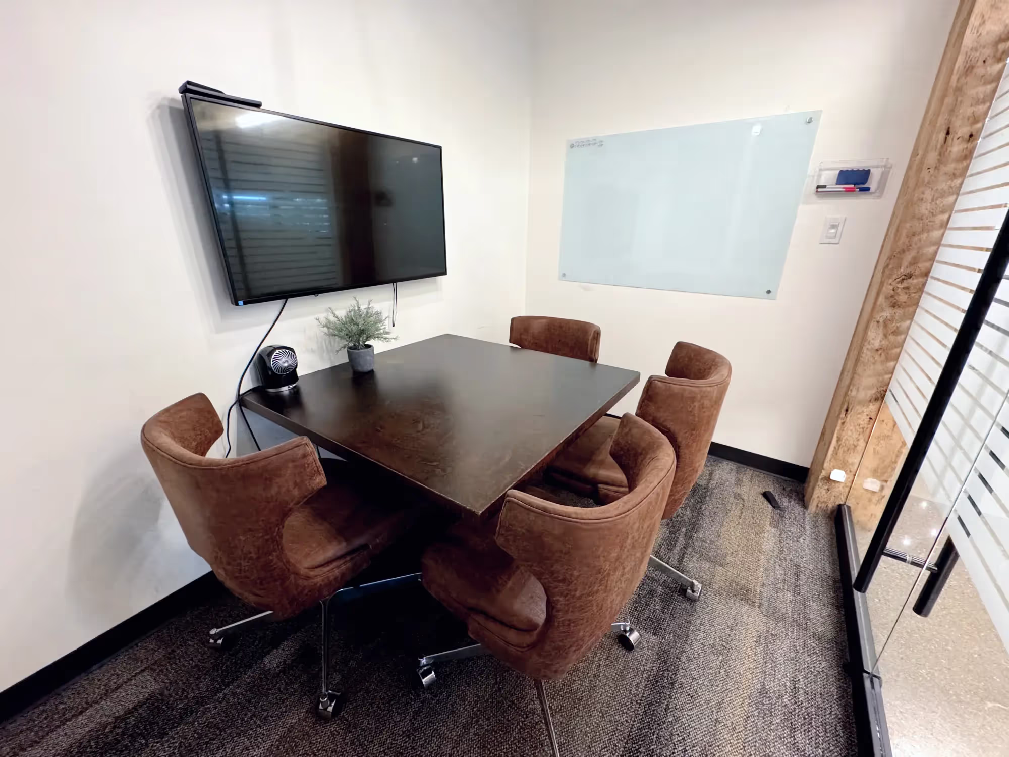 A small coworking space meeting room with a dark rectangular table, four brown swivel chairs, a wall-mounted TV, a whiteboard, a potted plant, and glass walls with frosted stripes.