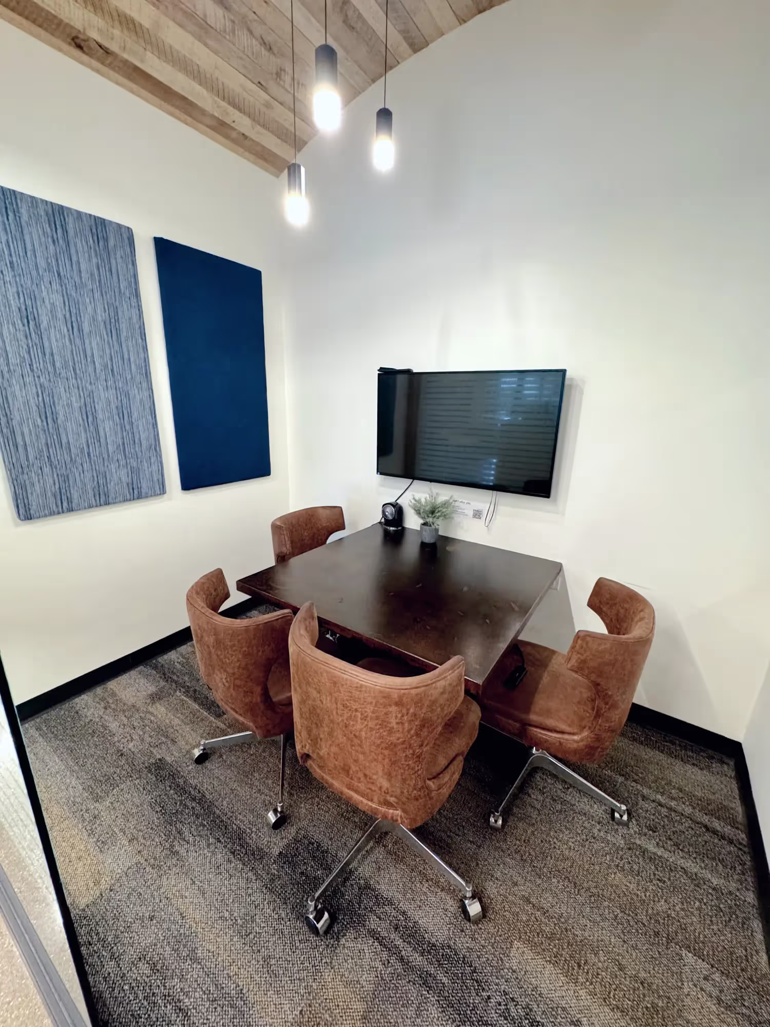Small modern meeting room in a coworking space, featuring a dark square table, four brown swivel chairs, wall-mounted TV, plant, two blue acoustic panels on one wall, and three pendant lights hanging from a wooden ceiling.