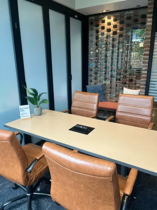 Modern office meeting room with a light wood table, four brown leather chairs, and a small plant. Ideal as an office space for rent or coworking space, with a glass wall, brick accent wall, and a cozy bench in the background.