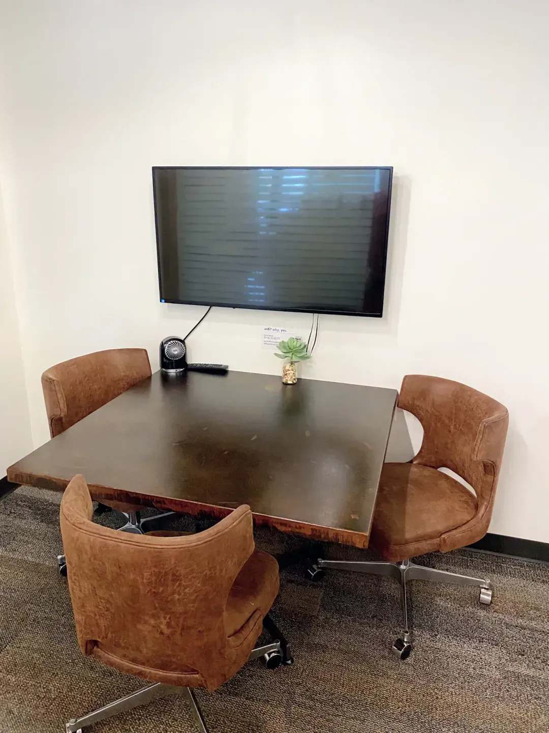 A small meeting room in an office space for rent with a dark square table, three brown swivel chairs, a wall-mounted TV, a small clock, and a potted succulent. The room features white walls and a carpeted floor—ideal for productive meetings.