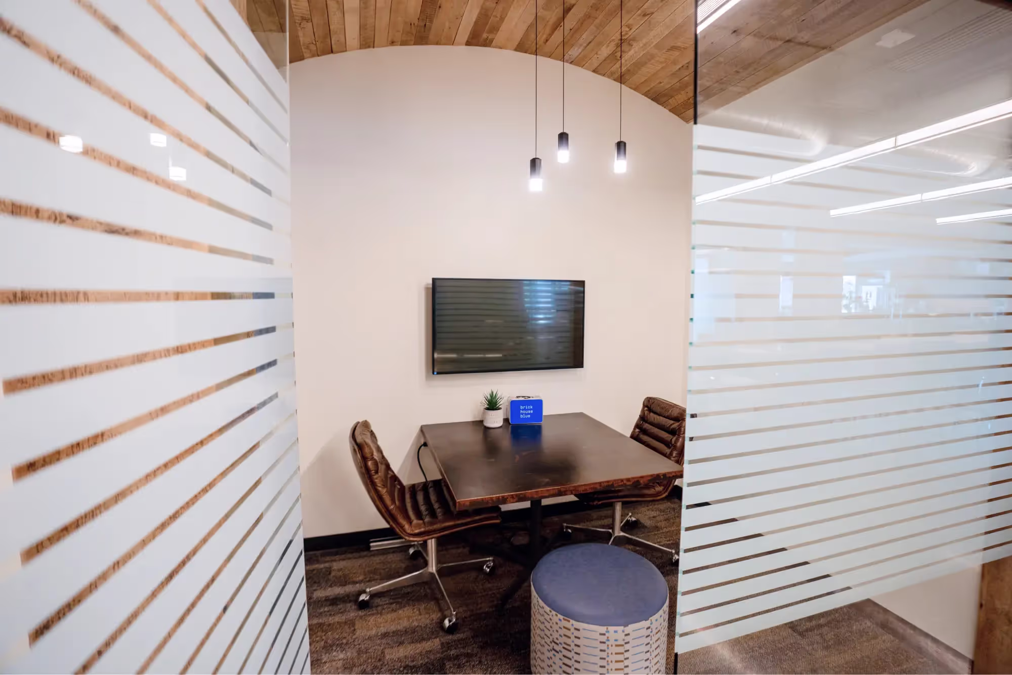 A small modern meeting room with a dark rectangular table, two brown chairs, a round blue stool, and a wall-mounted TV. Enclosed by glass walls with frosted stripes, it's ideal for meetings or as part of an office space for rent.