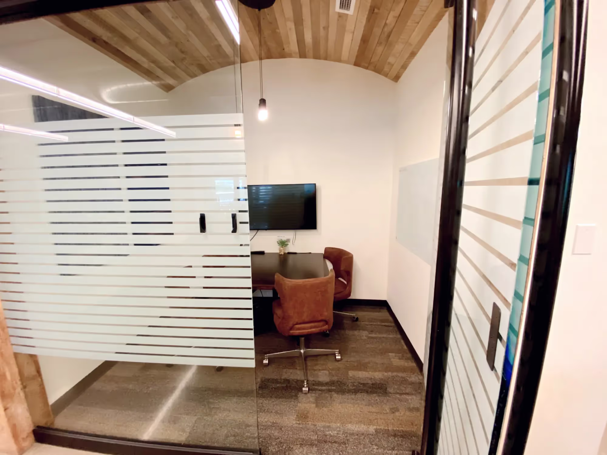 Small modern coworking space with glass walls, two brown chairs, a black table, a TV on the wall, a whiteboard, and a light wood ceiling. A single pendant light hangs above—ideal for private offices or meeting rooms.