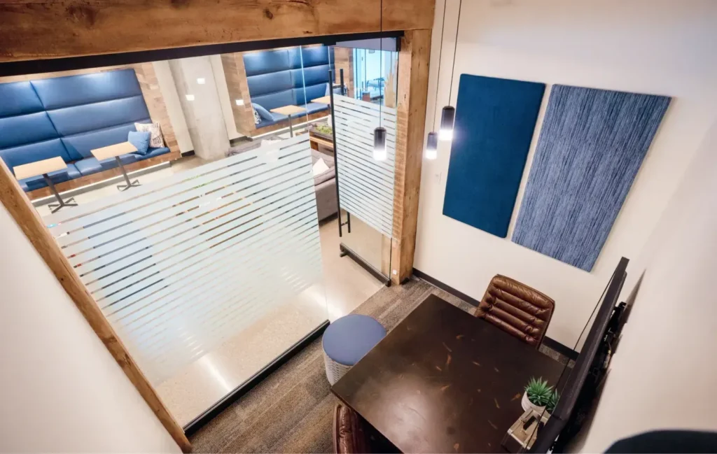 A modern coworking space viewed from above, featuring a dark wood desk with a brown leather chair, blue wall panels, frosted glass walls with horizontal stripes, and a glimpse of private offices and booths in the lounge area outside.