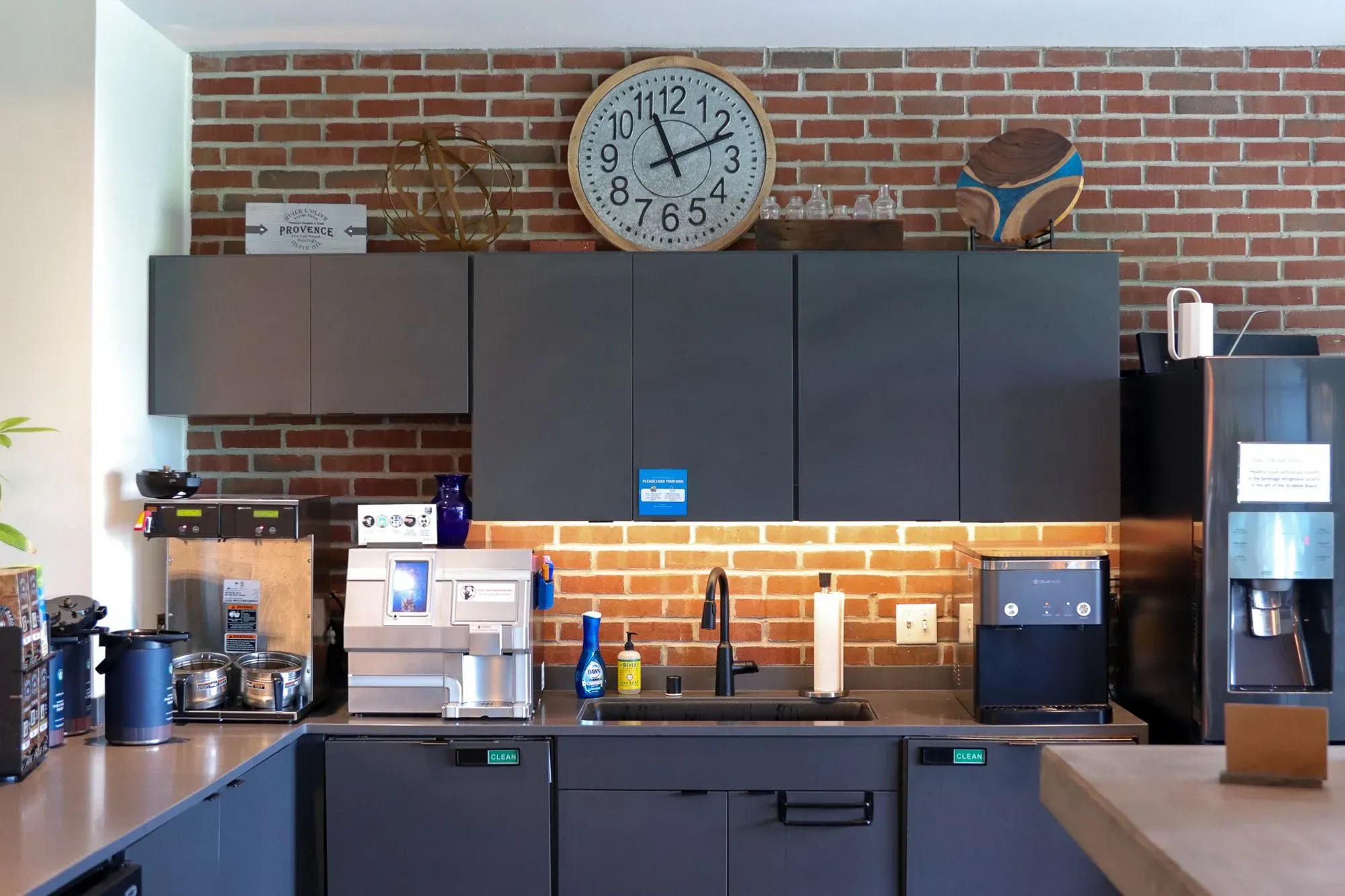Modern office kitchen in a coworking space features dark cabinets, coffee machines, and a sink beneath a large wall clock. The brick backsplash and organized items create a clean workspace, perfect for those seeking office space for rent. Warm lighting brightens the counter.
