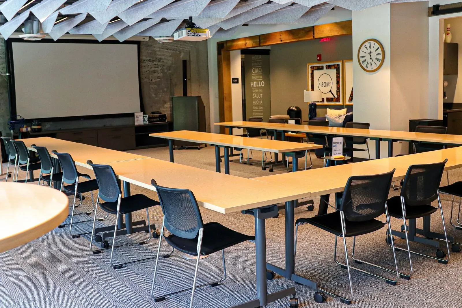A modern conference room in one of the top event spaces Columbus Ohio offers—featuring U-shaped tables, black chairs, a projector screen, and exposed ceiling. The well-lit space is ideal for meetings or those needing a virtual business address.