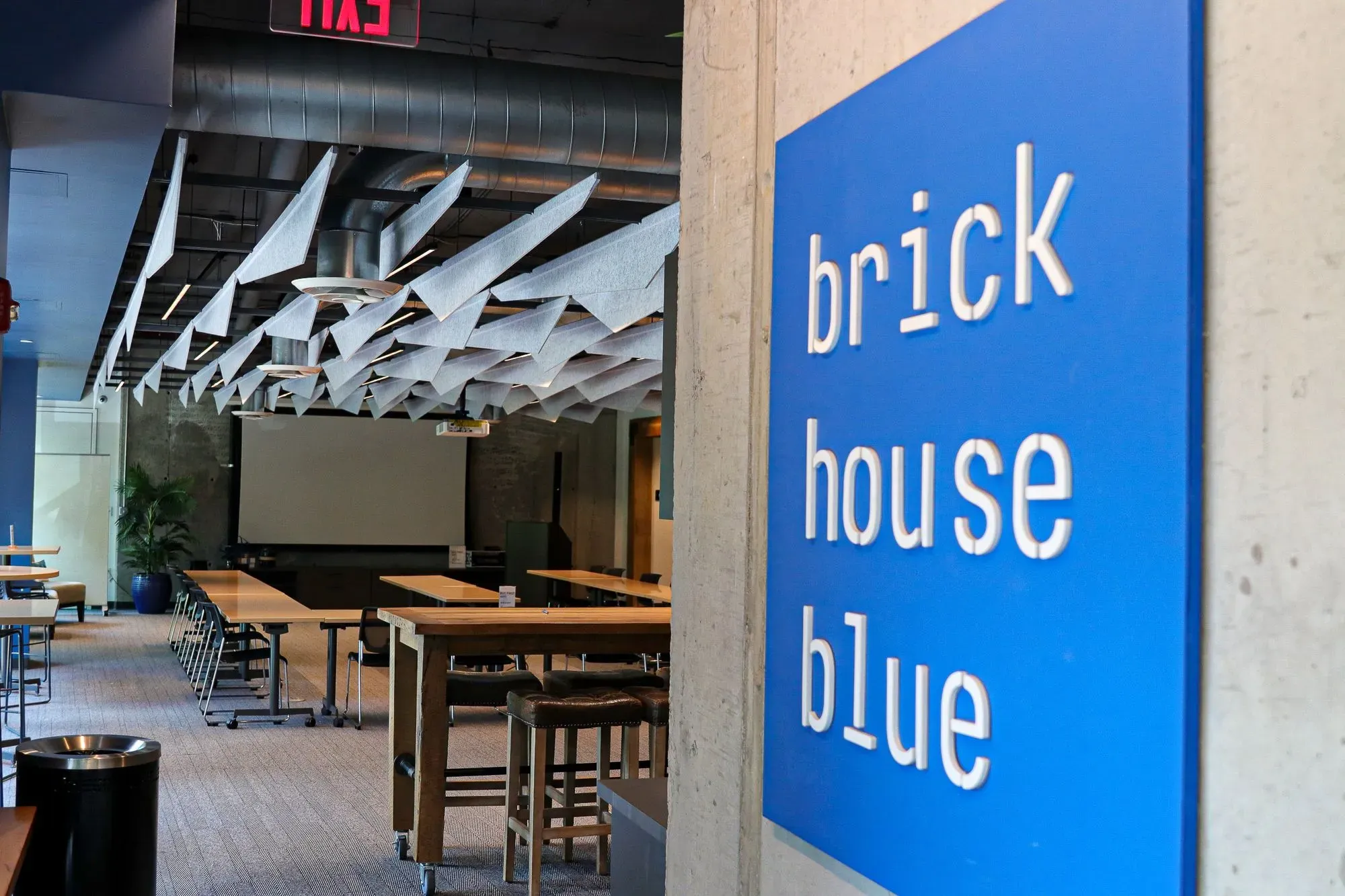 A blue sign with white text reads brick house blue on a concrete column inside a modern open workspace with tables, chairs, and geometric ceiling panels—ideal for those seeking office space for rent. An exit sign is visible in the background.