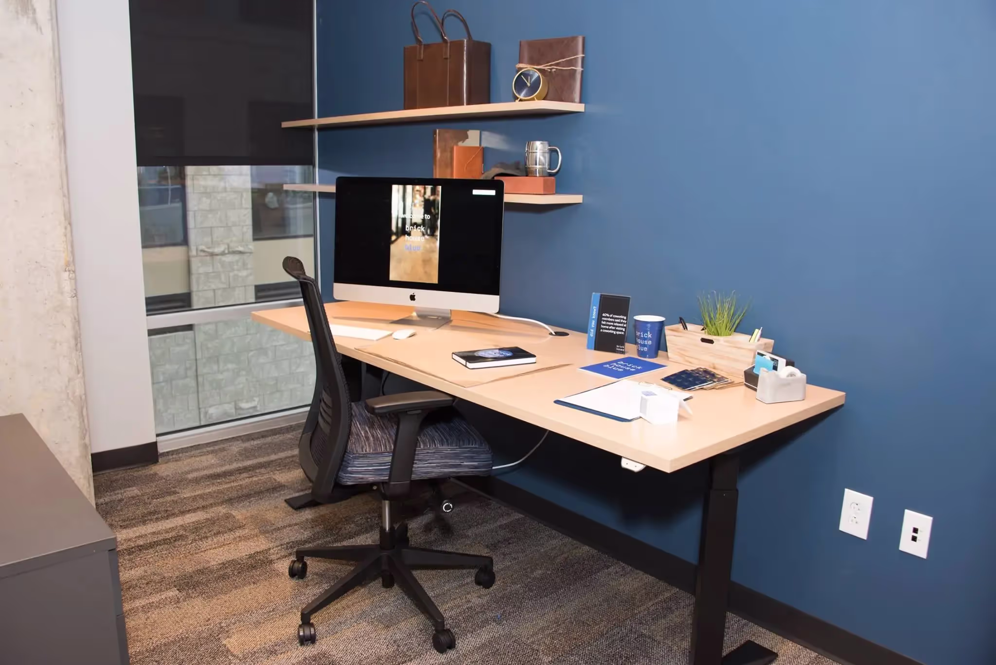A modern office workspace with a wooden desk, computer, office chair, notebooks, and plant. Floating shelves with decor are mounted on a blue wall beside a large window—ideal for those needing private offices or meeting rooms.