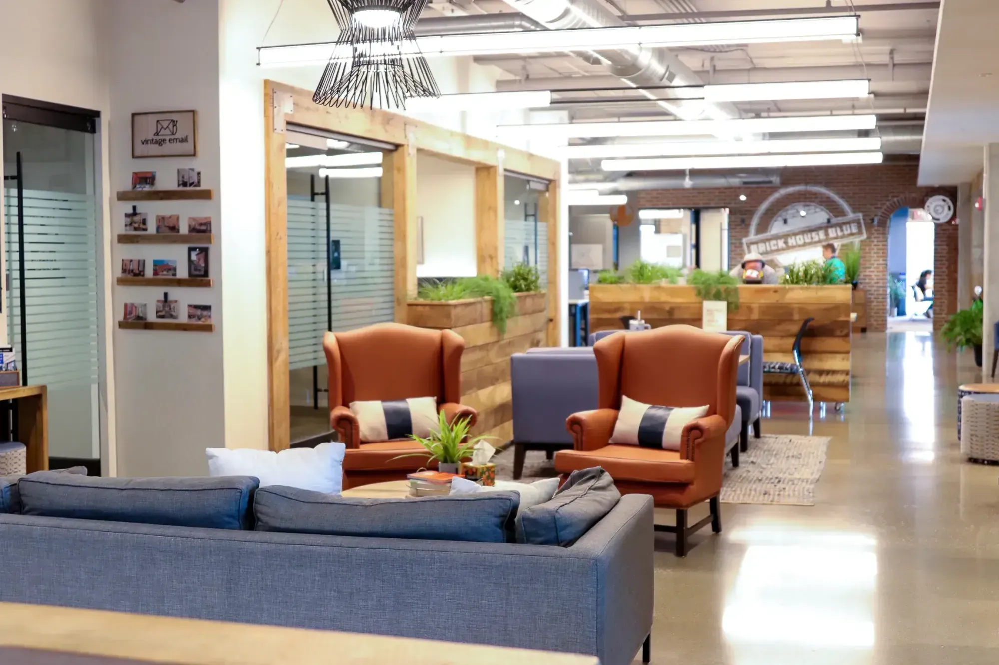 A modern office lounge with orange armchairs, a gray sofa, potted plants, and wooden partitions. Bright lighting and large windows create an open, welcoming atmosphere—perfect for networking or relaxing between meetings in private offices.
