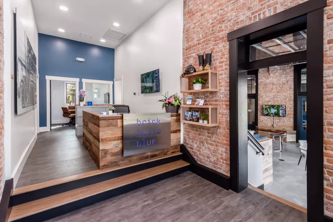 A modern office lobby with exposed brick walls, a wooden reception desk labeled brick house blue, wall shelves with decor, and a view into a stylish coworking space with windows, artwork, and flexible meeting rooms.