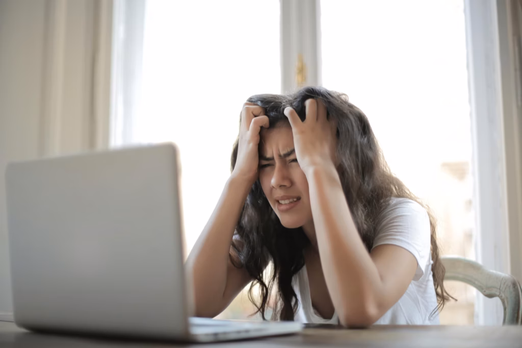 A woman with long dark hair sits at a table in front of a laptop, holding her head in frustration and stress—perhaps overwhelmed by searching for virtual business address options or meeting rooms. Bright daylight shines through the window behind her.