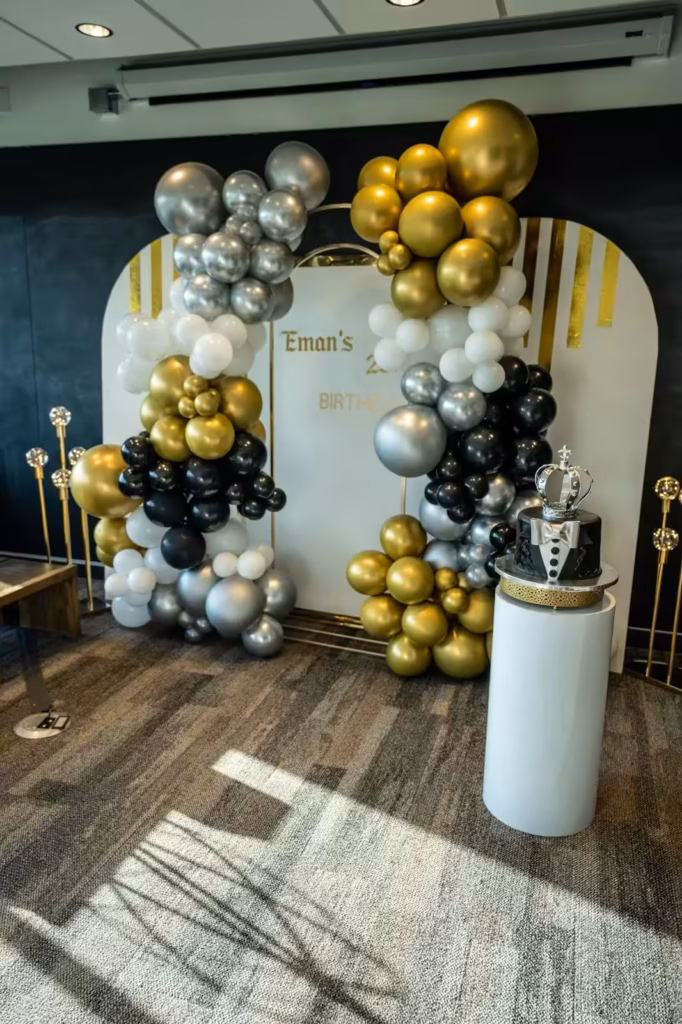 A birthday party setup with a gold, silver, black, and white balloon arch around a sign reading Emans Birthday, plus a black and gold cake topped with a crown on a white pedestal—perfect inspiration for your next coworking space celebration.