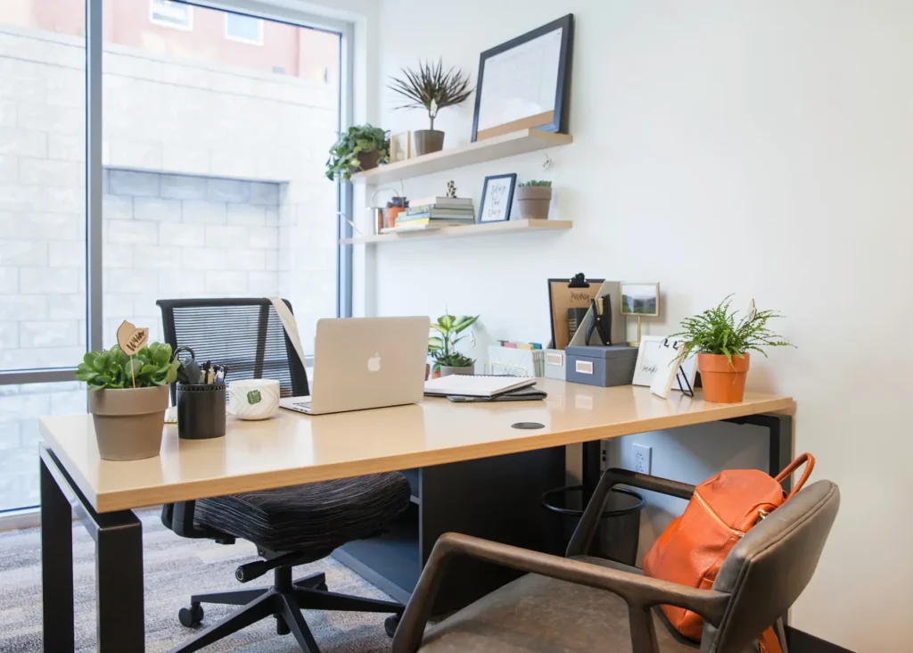 A modern coworking space with a wooden desk, laptop, plants, office supplies, and shelves with books and decor. A window lets in natural light, and a brown chair with an orange bag is in the foreground.