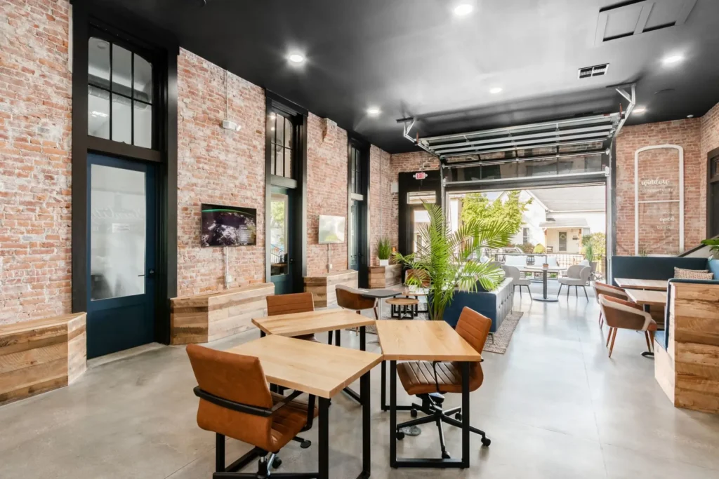 Modern open office with exposed brick walls, wood desks and chairs, indoor plants, and large windows providing natural light. Includes private offices, and a garage-style door opens to an outdoor patio.