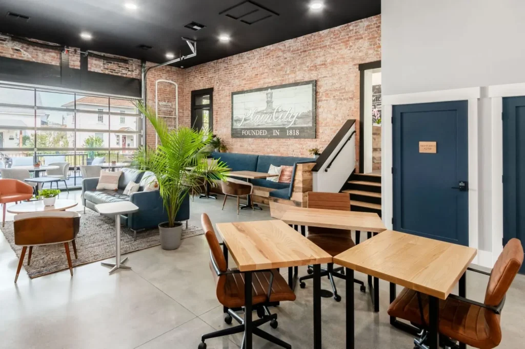 Modern coworking space with wooden tables, brown chairs, a potted plant, and a lounge area with sofas. Exposed brick walls, large windows, and a sign that reads “Salt City Founded in 1818” enhance this top choice for private offices in Columbus, Ohio.