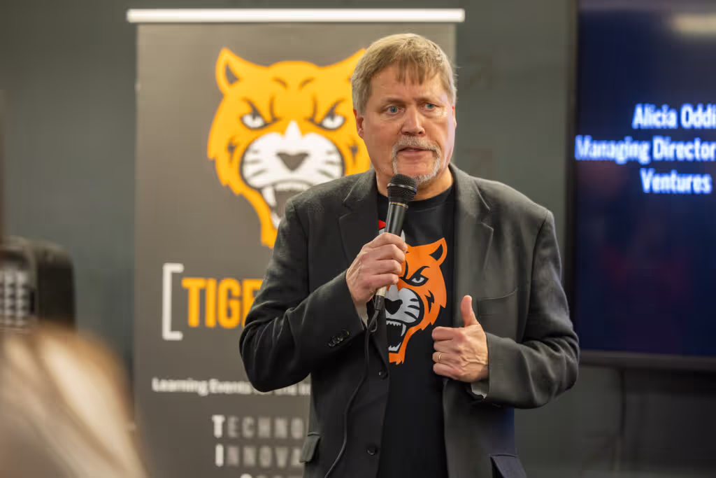 A man with a mustache and beard speaks into a microphone. He wears a black jacket and a shirt featuring an orange tiger face, standing in front of a banner with a large tiger illustration at a coworking space.