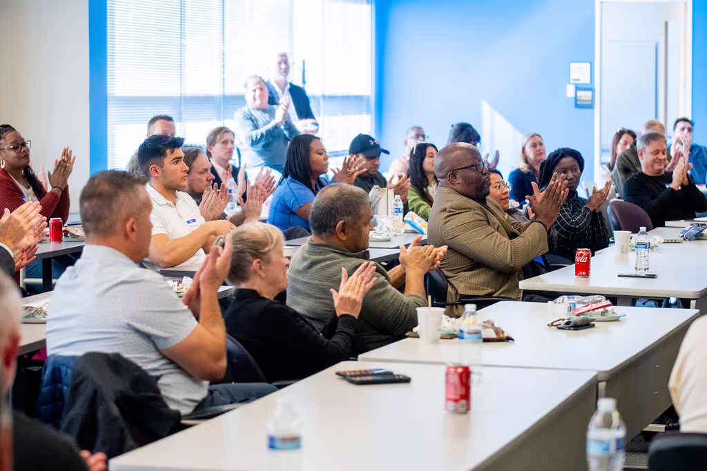 A diverse group sits at tables in a brightly lit room, clapping and engaged with a speaker. Snacks and drinks are on the tables, creating a positive atmosphere—an ideal example of vibrant event spaces Columbus Ohio has to offer.
