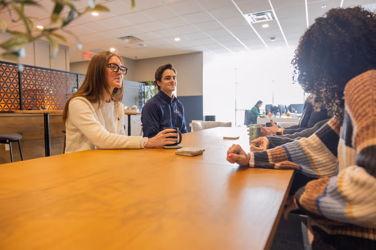 Four people sit around a wooden table in a bright, modern coworking space, engaged in conversation. Two face the camera, while the others are seen from behind. One person holds a mug, and notebooks are on the table.