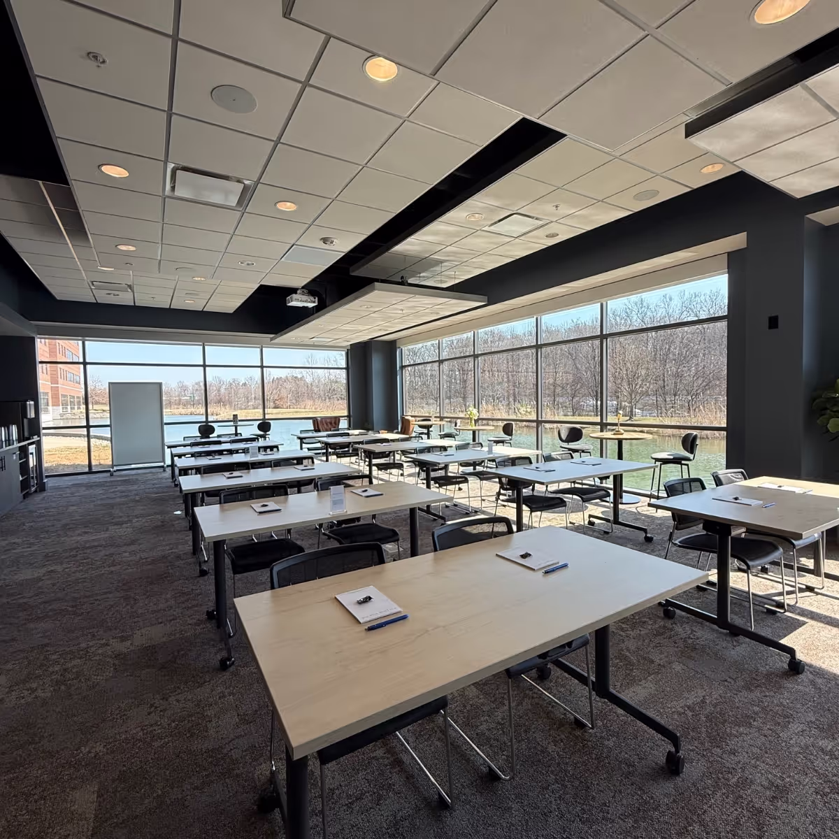 A modern conference room in one of the top event spaces Columbus Ohio offers, with large windows, rows of tables and chairs, and notepads with pens on each table. Sunlight pours in, revealing a grassy outdoor area with trees just outside.