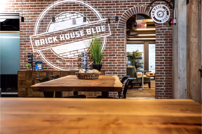 A modern coworking space with exposed brick walls, wooden tables, greenery, and a large Brick House Blue logo projected on the wall. An archway leads to more seating, lounge chairs, and flexible meeting rooms filled with plants.