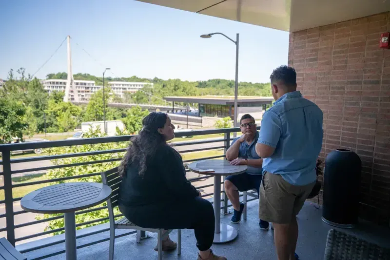 Three people are talking on a balcony with round tables and chairs at a coworking space. Trees, buildings, and a street are visible in the background on a sunny day.