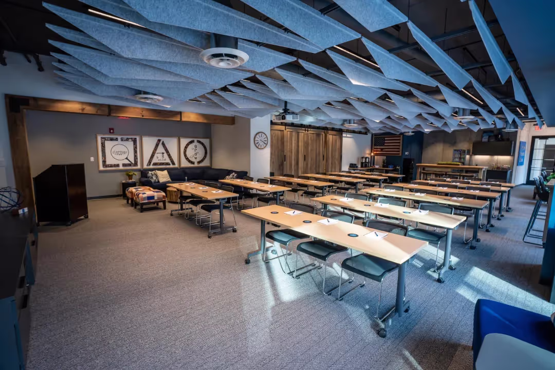 A modern classroom with wooden tables and chairs arranged in rows, acoustic panels on the ceiling, and various decorations on the walls—including framed prints, clocks, an American flag, and a corner area styled to resemble private offices.