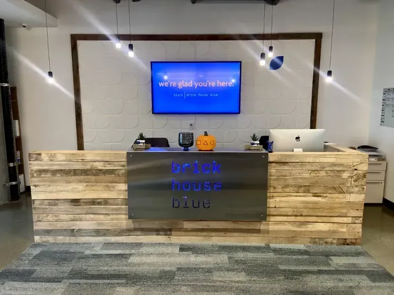 A modern reception desk made of light wood with a metal sign reading brick house blue. Behind the desk is a TV displaying a welcome message and shelves with office supplies and small plants—perfect for a coworking space in Columbus, Ohio.