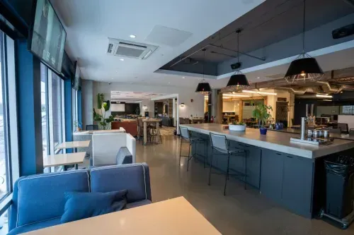 A modern, spacious coworking space with large windows, blue cushioned seating, high-top tables, pendant lights, and a polished floor, creating a bright and inviting atmosphere.