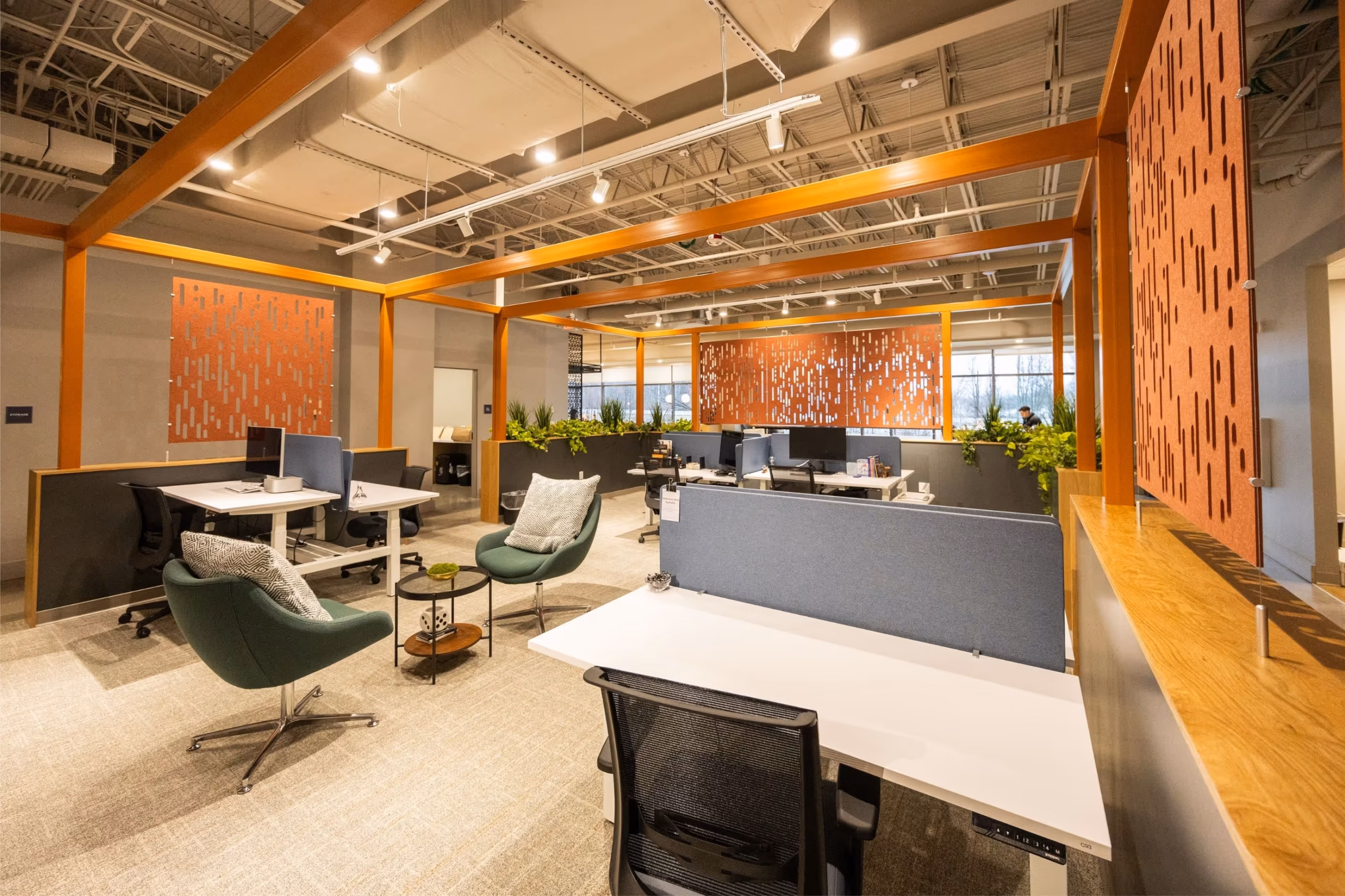 A modern open-plan coworking space with desks, ergonomic chairs, partitions, green plants, and warm lighting. Decorative orange panels hang above workstations, while large windows fill the clean and inviting space with natural light.