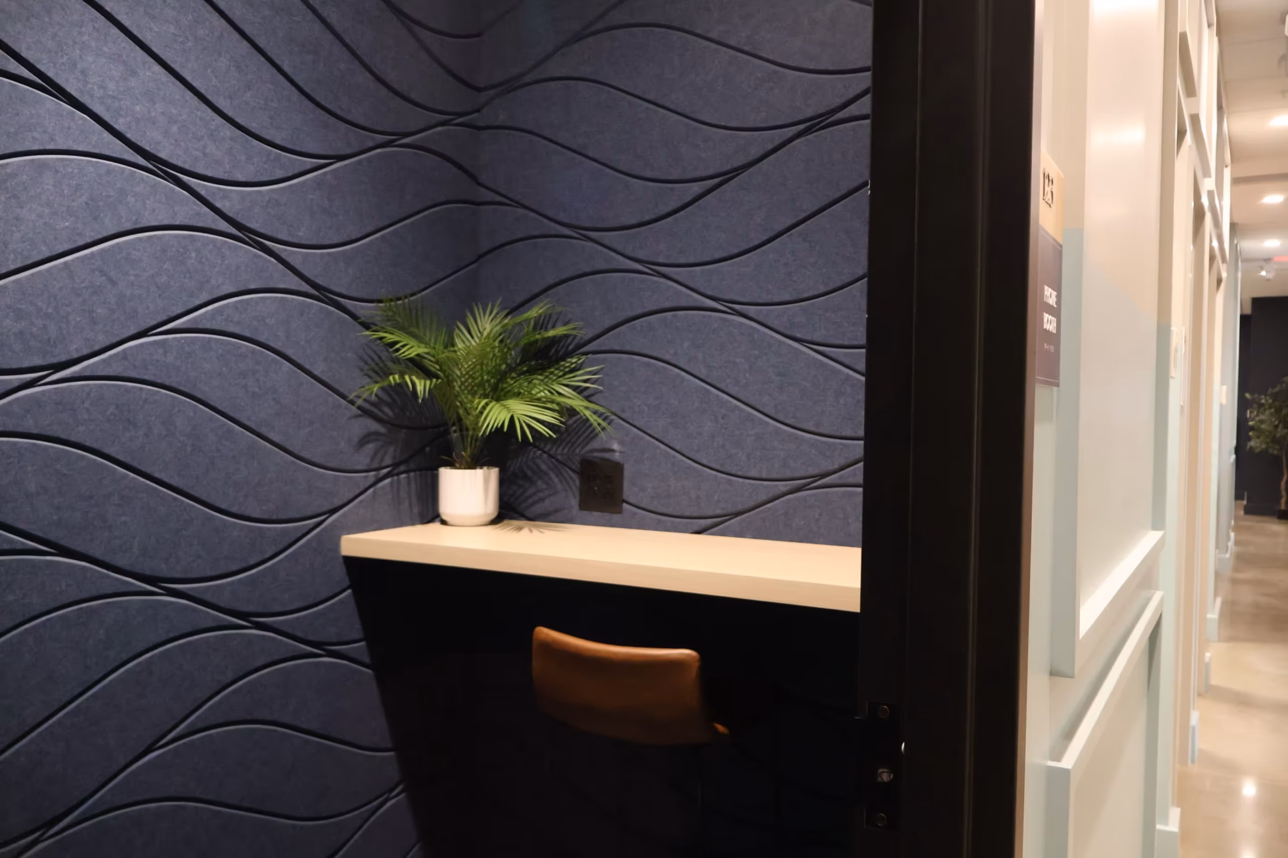 A small modern office nook with a dark wavy textured wall, a potted green plant on a beige counter, a brown chair, and access to meeting rooms just beyond the open door—ideal for productivity and collaboration.
