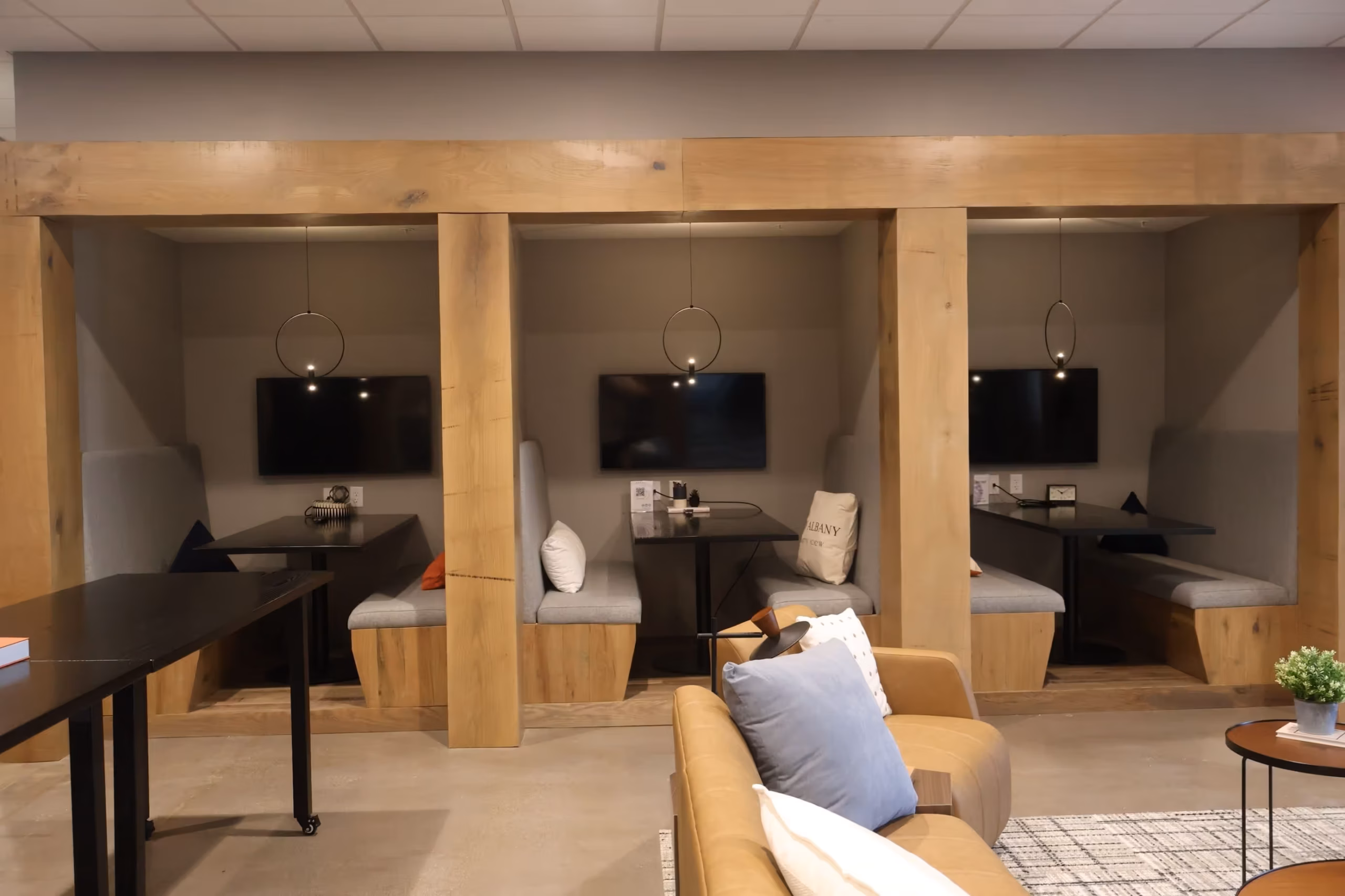 Three modern wooden booths with gray cushioned seating and black tables, each with a TV screen and pendant light, are built into a wall. In front, a tan sofa and small black table with a potted plant create an inviting event space in Columbus, Ohio.