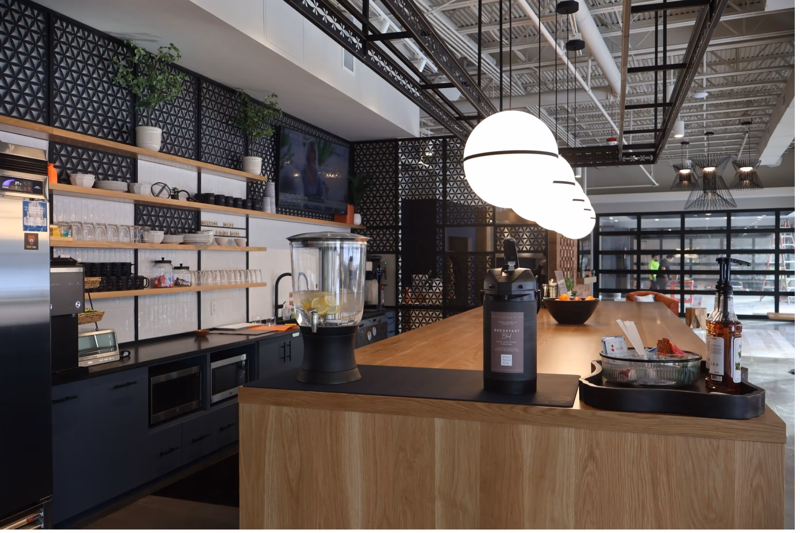 A modern kitchen with a large wooden island, beverage dispenser, hanging round lights, open shelves with dishes, black cabinets, and decorative wall panels—perfect for casual gatherings or event spaces in Columbus, Ohio. Snacks and drinks are set on the counter.