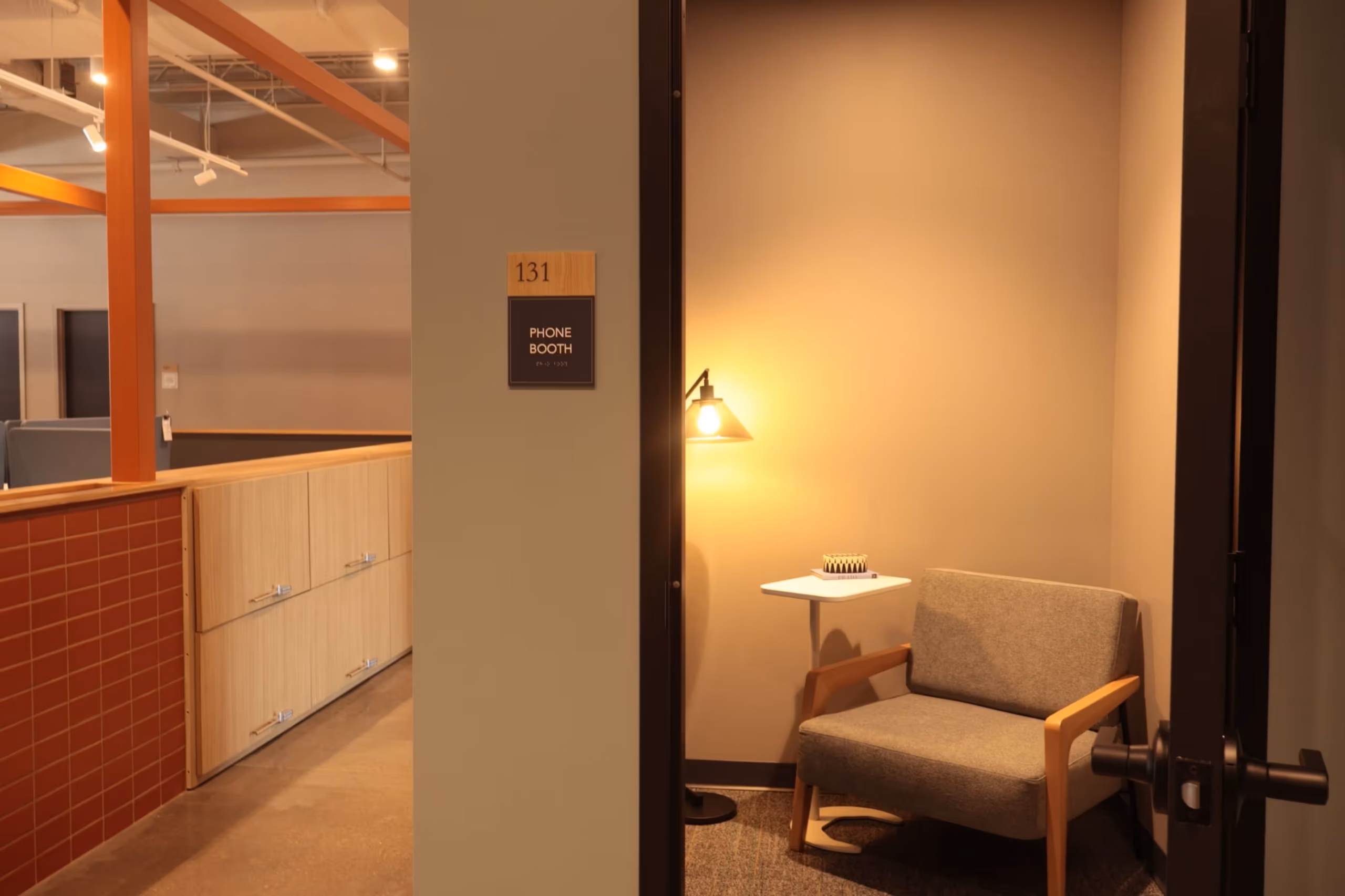 A small office phone booth with a gray armchair, a white side table, a lamp, and a chess set. Labeled 131 Phone Booth, it opens to modern office space with wood and brick finishes—perfect for meeting rooms or event spaces Columbus Ohio.