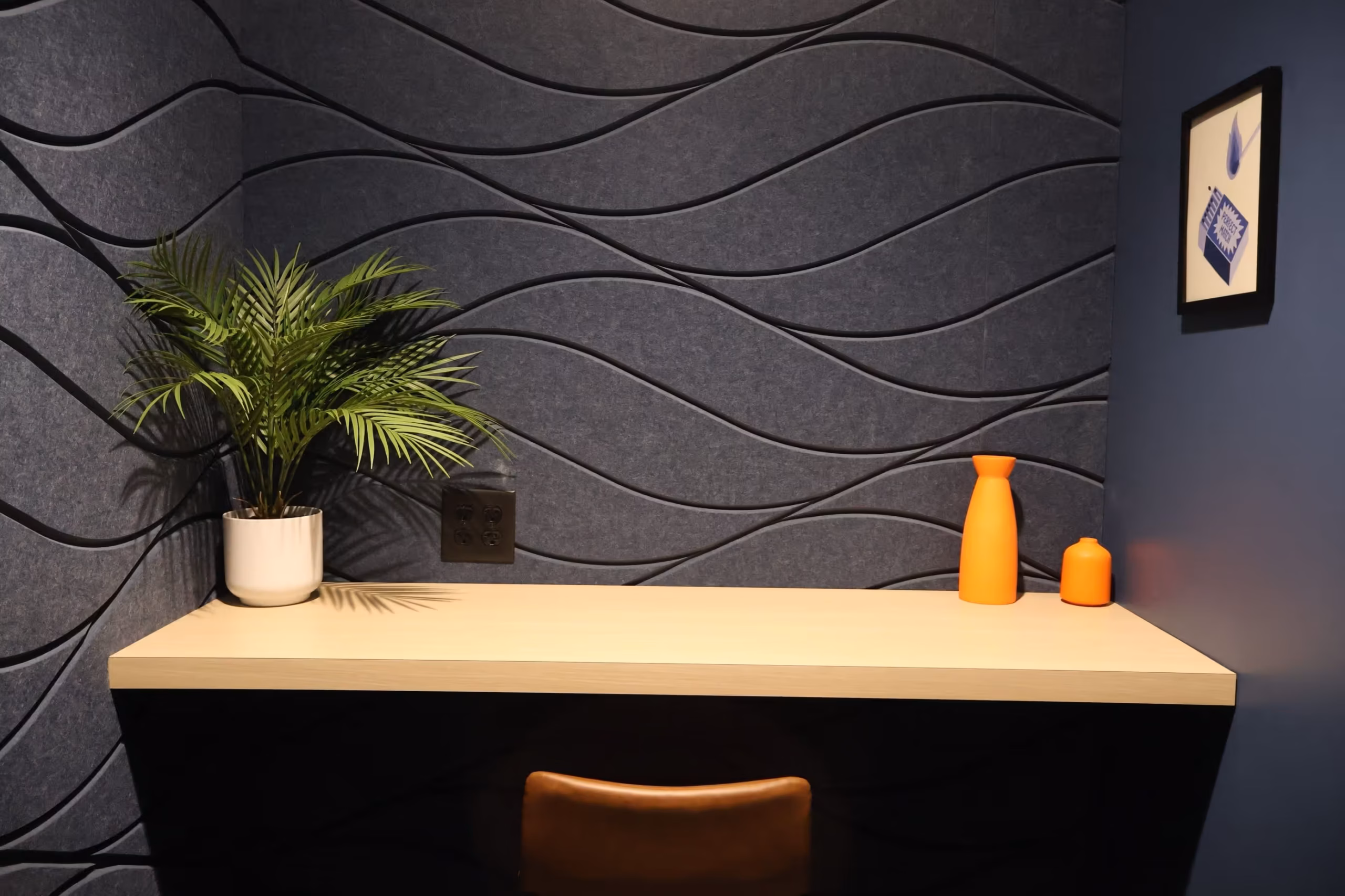 A minimalist coworking space desk with a brown chair, potted plant, orange vase, and small orange object. The wall features a dark blue wavy pattern with a framed picture in the top right corner.