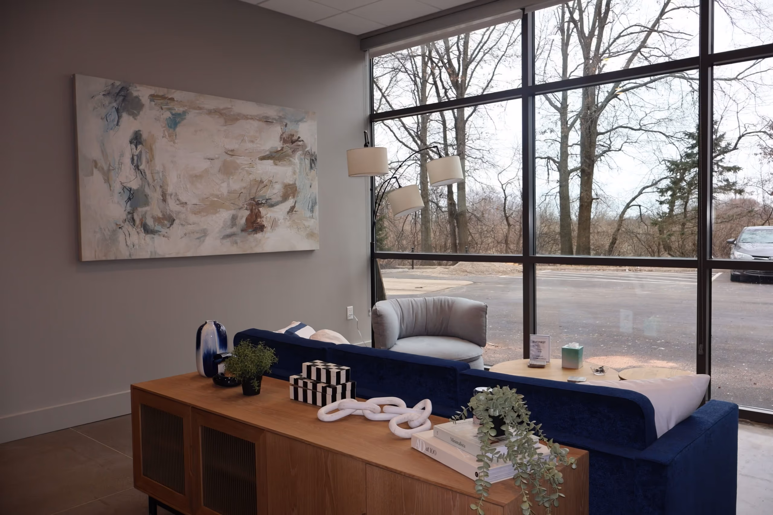 A modern lounge with a blue sofa, gray armchair, abstract wall art, and a wooden console table creates a welcoming coworking space. Large windows reveal leafless trees as natural light brightens the room, making it ideal for relaxation or meetings.