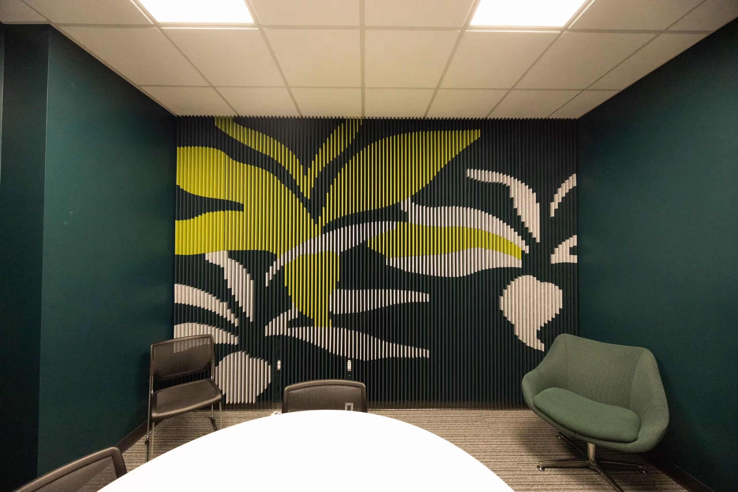 A modern office meeting room in a coworking space with dark green walls, a decorative wall featuring a large abstract leaf design, a round white table, several chairs, and a green upholstered lounge chair.
