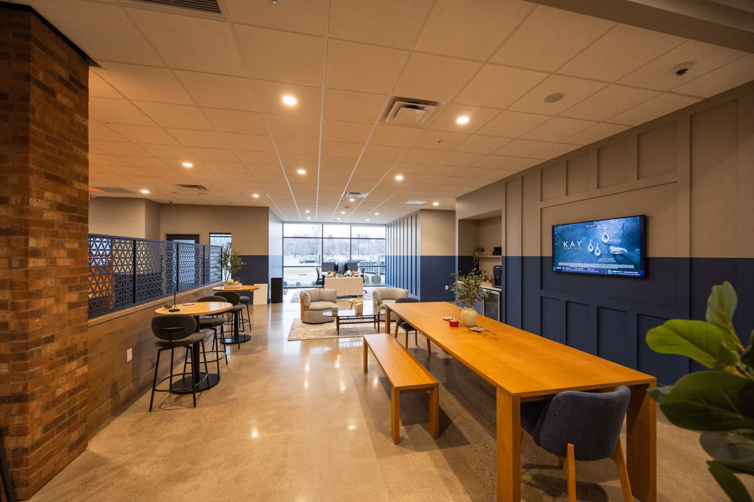 Modern lounge area with wooden tables and chairs, a TV on the wall, and sofas by large windows. Ideal office space for rent with natural light, plants, and subtle blue and brick accents to enhance productivity and comfort.
