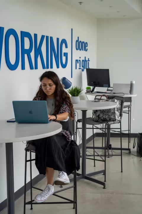 A woman with long hair sits at a high table, typing on a laptop in a modern coworking space. Behind her, blue letters on the wall read “WORKING done right.” Nearby, tall chairs and desks hold bags and office supplies.