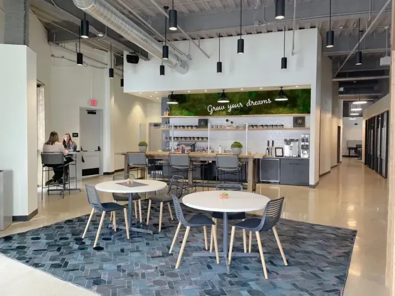 Modern coworking space lounge with round tables and chairs on a blue rug, a coffee bar in the background, and a neon “Grow your dreams” sign. Two people chat near the counter—perfect for meetings or office space for rent.