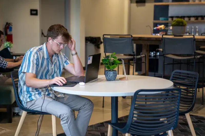 A young man wearing glasses and earphones works on a laptop at a round table in a modern coworking space. There is a small potted plant on the table and high chairs in the background.