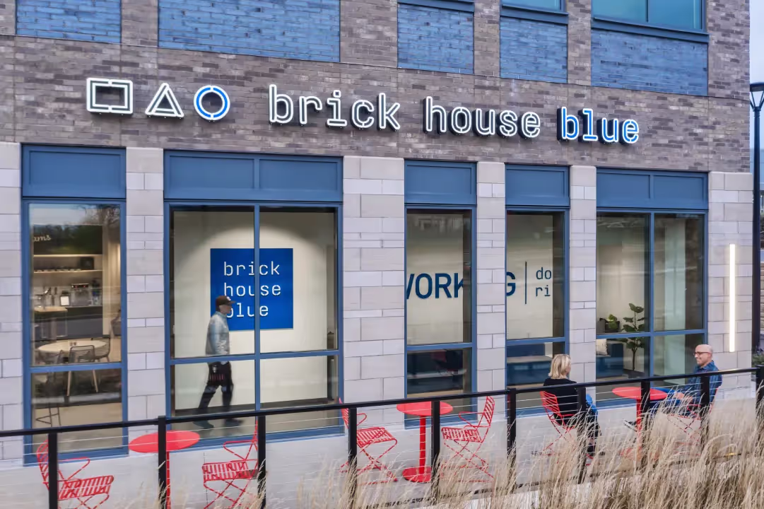 A modern building facade with the sign brick house blue in neon, above large windows. Red outdoor tables and chairs line the walkway, with two people seated and another walking inside—an inviting coworking space and office space for rent.
