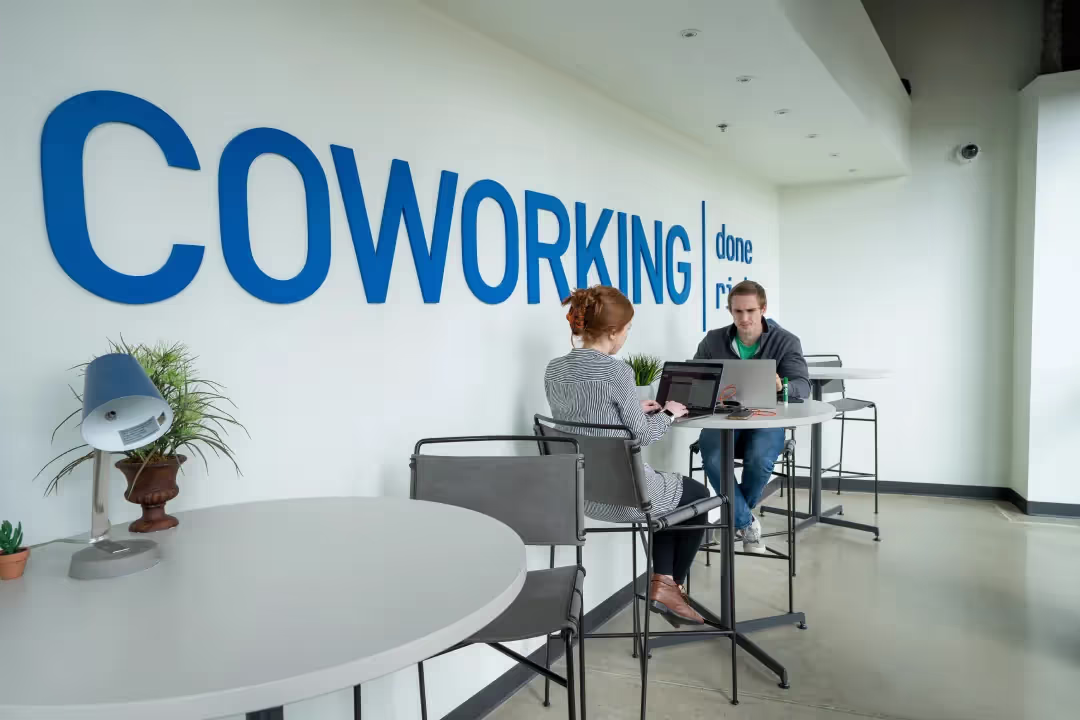 Two people work on laptops at high tables in a modern coworking space with large blue COWORKING done right text on the wall; this bright, minimalist area offers office space for rent and event spaces Columbus Ohio professionals love.