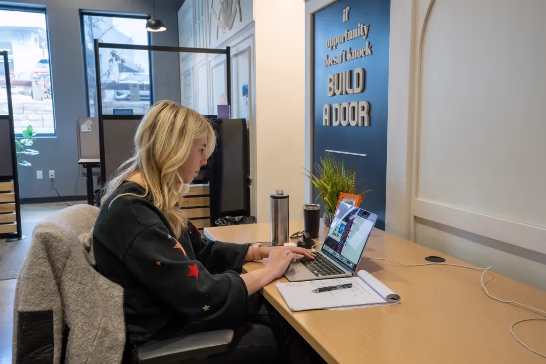 A woman with blonde hair is working on a laptop at a desk in a modern coworking space. There’s a notebook, pen, and water bottle on the desk. A sign on the wall reads, If opportunity doesn’t knock, build a door.