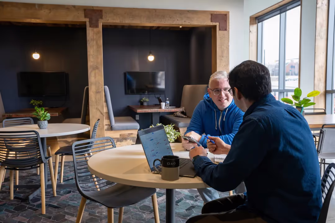 Two people sit at a round table in a modern coworking space, engaged in conversation. An open laptop and a mug rest on the table. Cozy booths, lush plants, and warm lighting in the background create an inviting office space for rent.