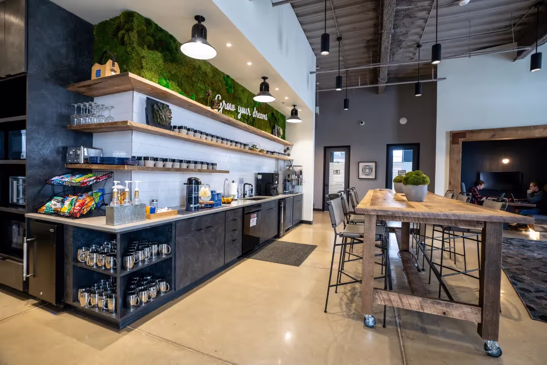 A modern coworking space kitchen with high ceilings, a long wooden table and chairs, a coffee and snack station, shelves with cups and plants, and a moss wall art feature. A lounge area with people is visible in the background.