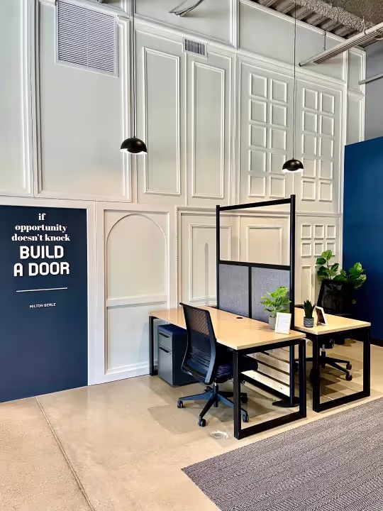 Modern office space for rent with two wooden desks, black chairs, a partition, and plants. A wall sign reads, If opportunity doesn't knock, build a door. Features white paneled walls, hanging black lights—ideal for meetings or event spaces in Columbus, Ohio.